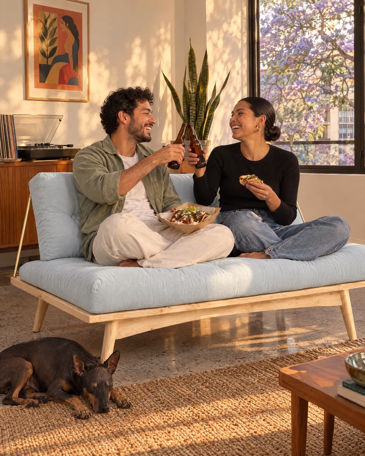 Dos amigos brindan con cerveza y tacos al pastor sentados en un Futon Folk color Cielo, en un departamento de la Condesa al atardecer